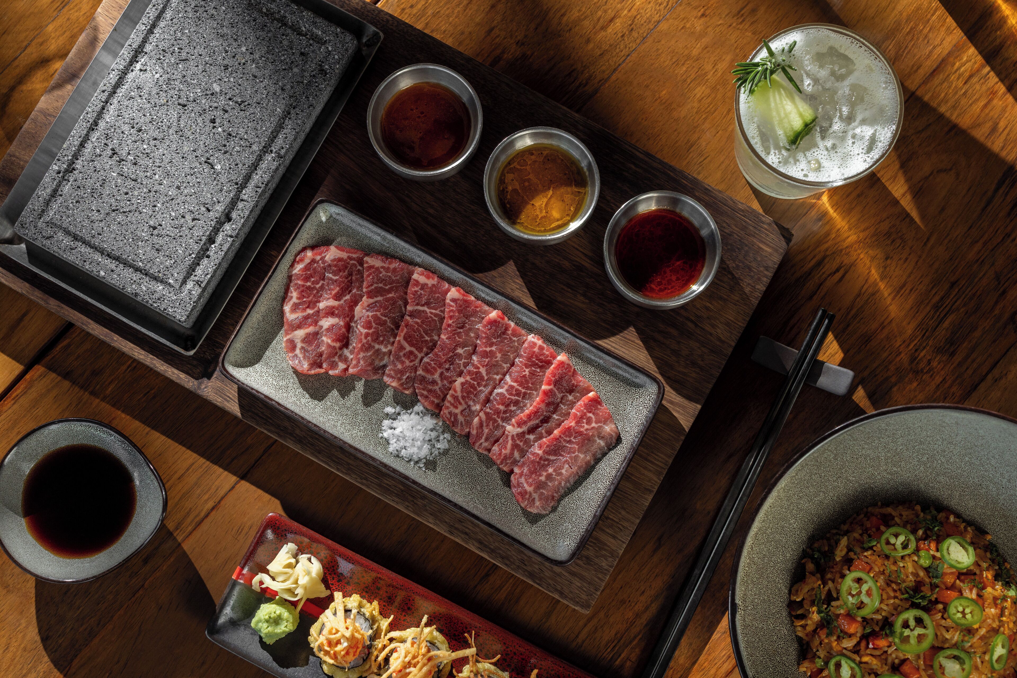 A dining table with a hot plate, raw meat, condiments, and a bowl of rice.