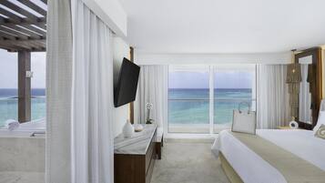 A hotel room with a large window, a bed, a TV, a bathroom with a bathtub, and a view of the ocean.
