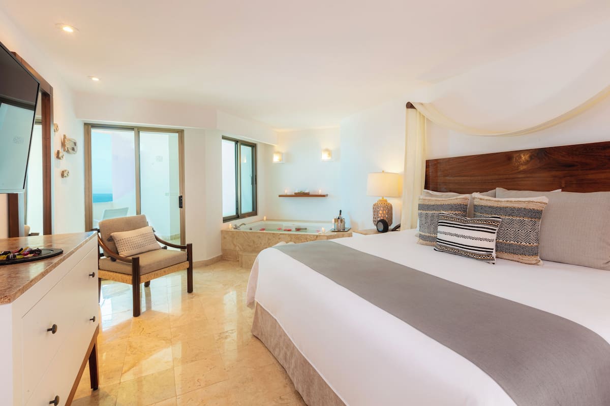 A modern hotel room with a large bed, a sitting area with a chair, a small table, a bathtub, and a TV.