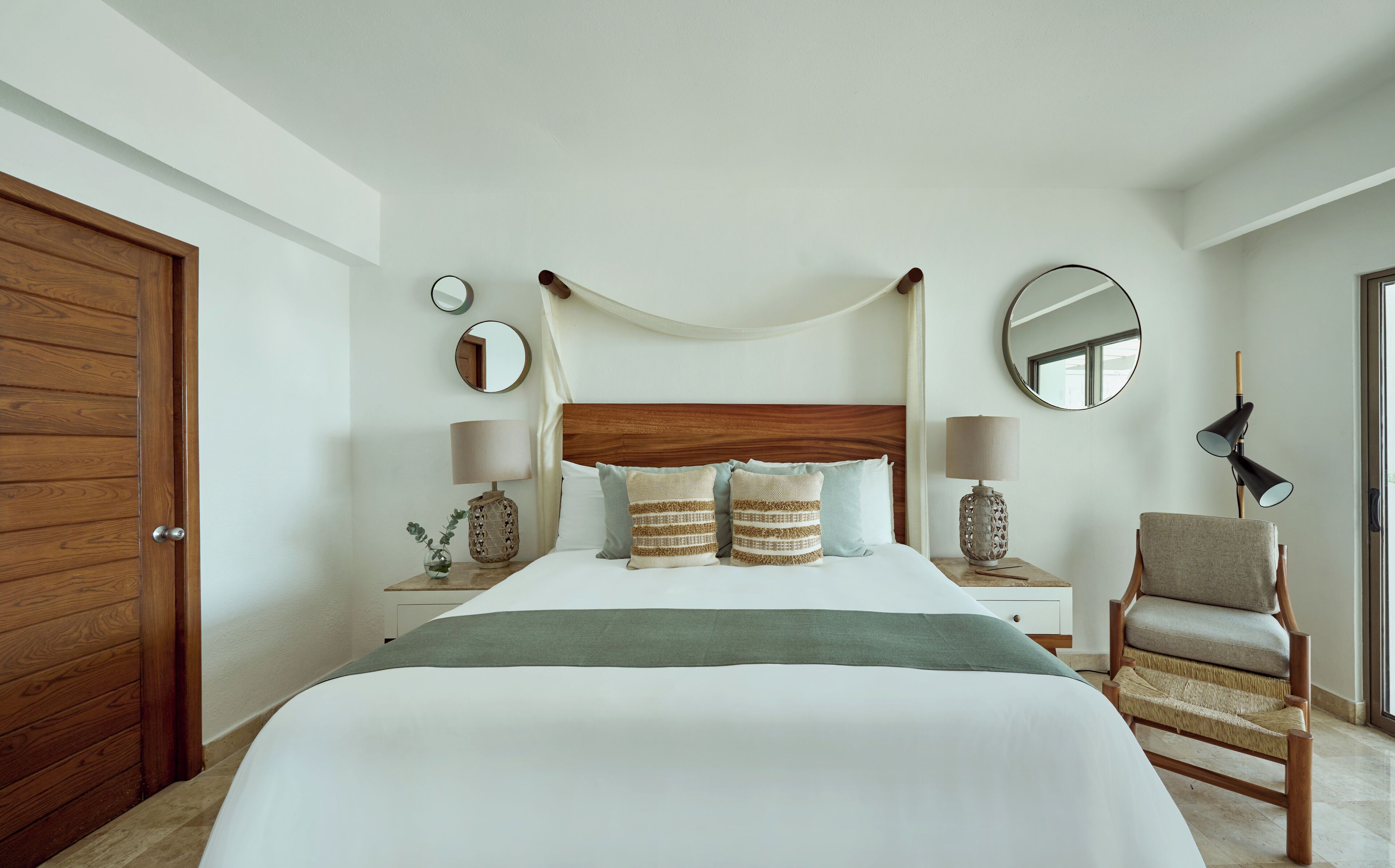 Un dormitorio ordenado con una cama grande, dos mesitas de noche con lámparas, una silla y una puerta de madera.