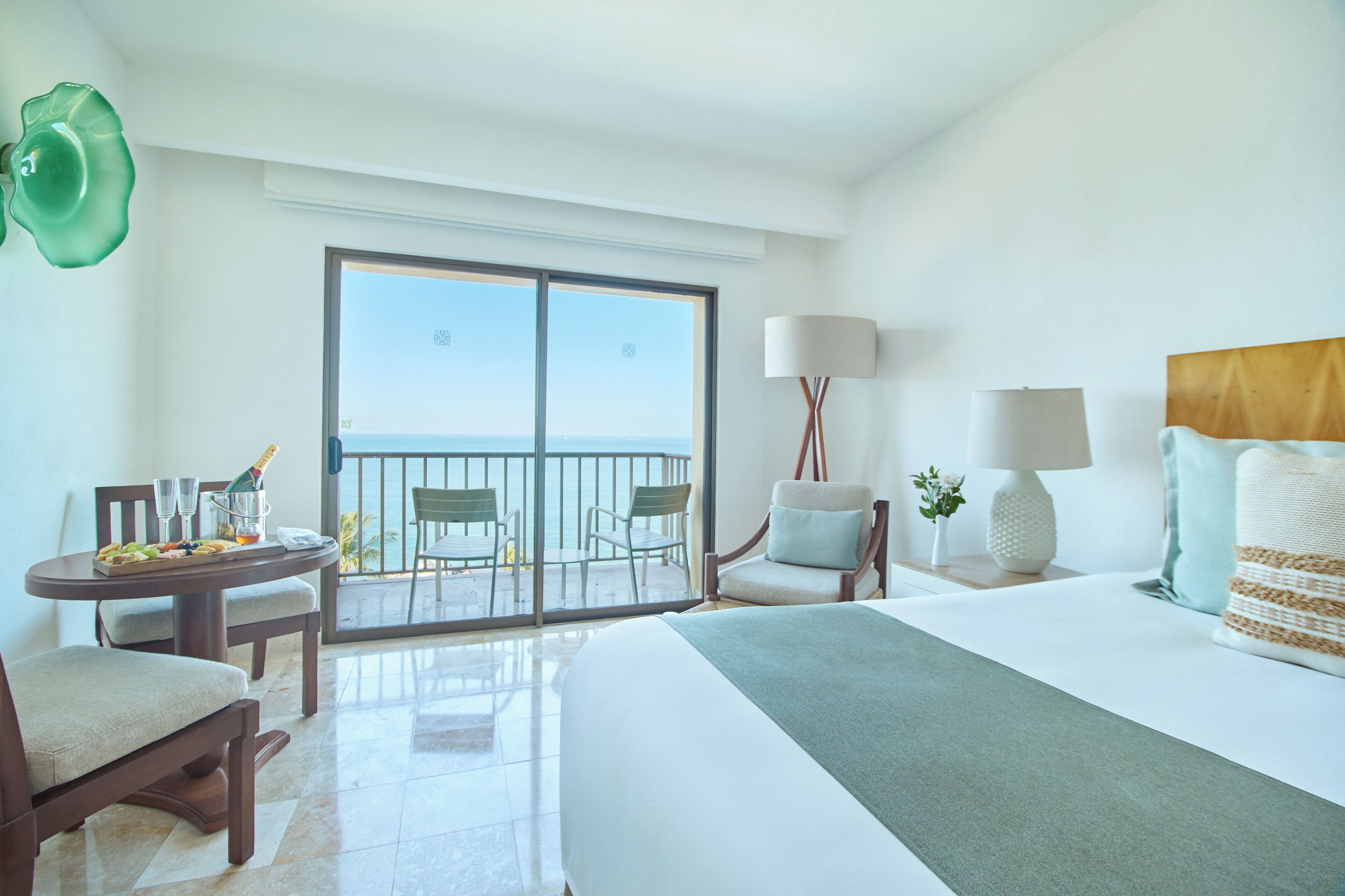 A hotel room with a bed, a dining table with chairs, a balcony with a view of the ocean, and a lamp.