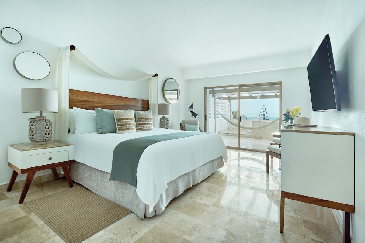 A modern hotel room with a bed, bedside tables, a desk, a TV, and a view of the sea through a glass door.