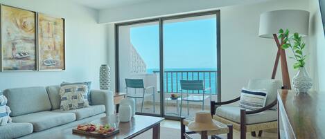 A modern living room with a sofa, coffee table, and chairs, a balcony with a view of the sea, and artwork on the wall.