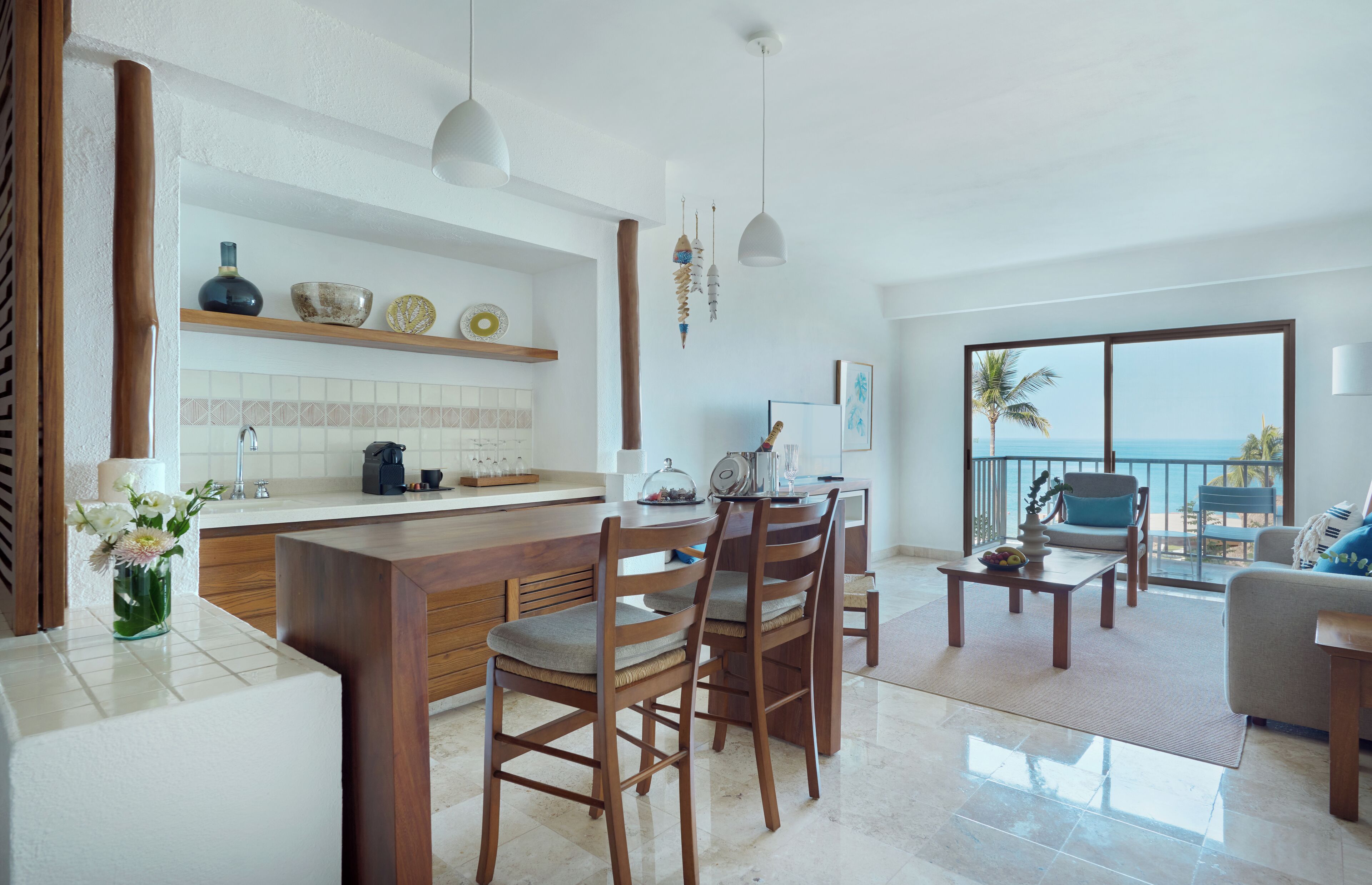 Une cuisine moderne avec un îlot central, des chaises en bois, une table à manger et une vue sur l’océan grâce à de grandes fenêtres.