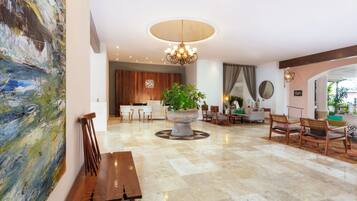 Um hall amplo com uma grande pintura abstrata, uma planta em vaso central, um candelabro e um balcão de recepção.