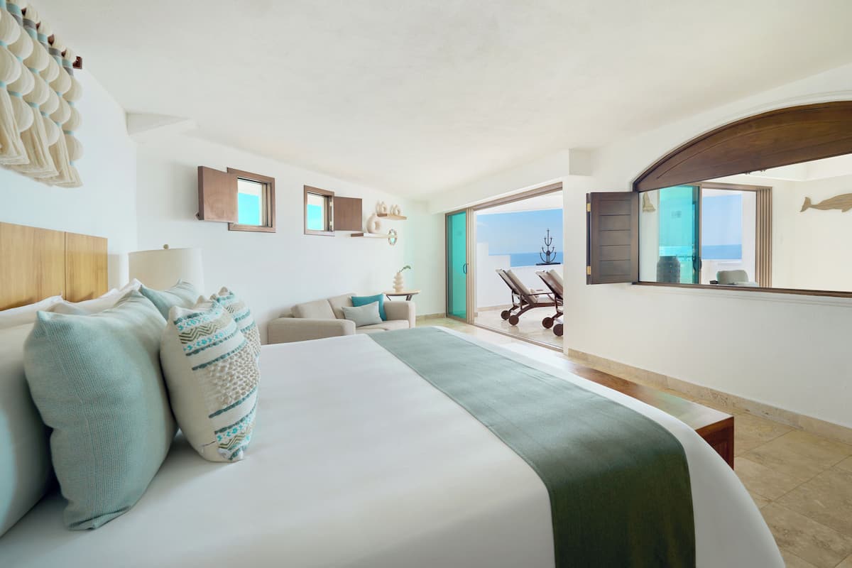 A hotel room with a large bed, a sofa, a dining table, and a view of the sea through a glass door.