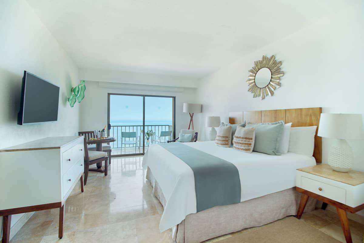 A hotel room with a bed, bedside tables, a desk, a TV, and a balcony with a view of the sea.