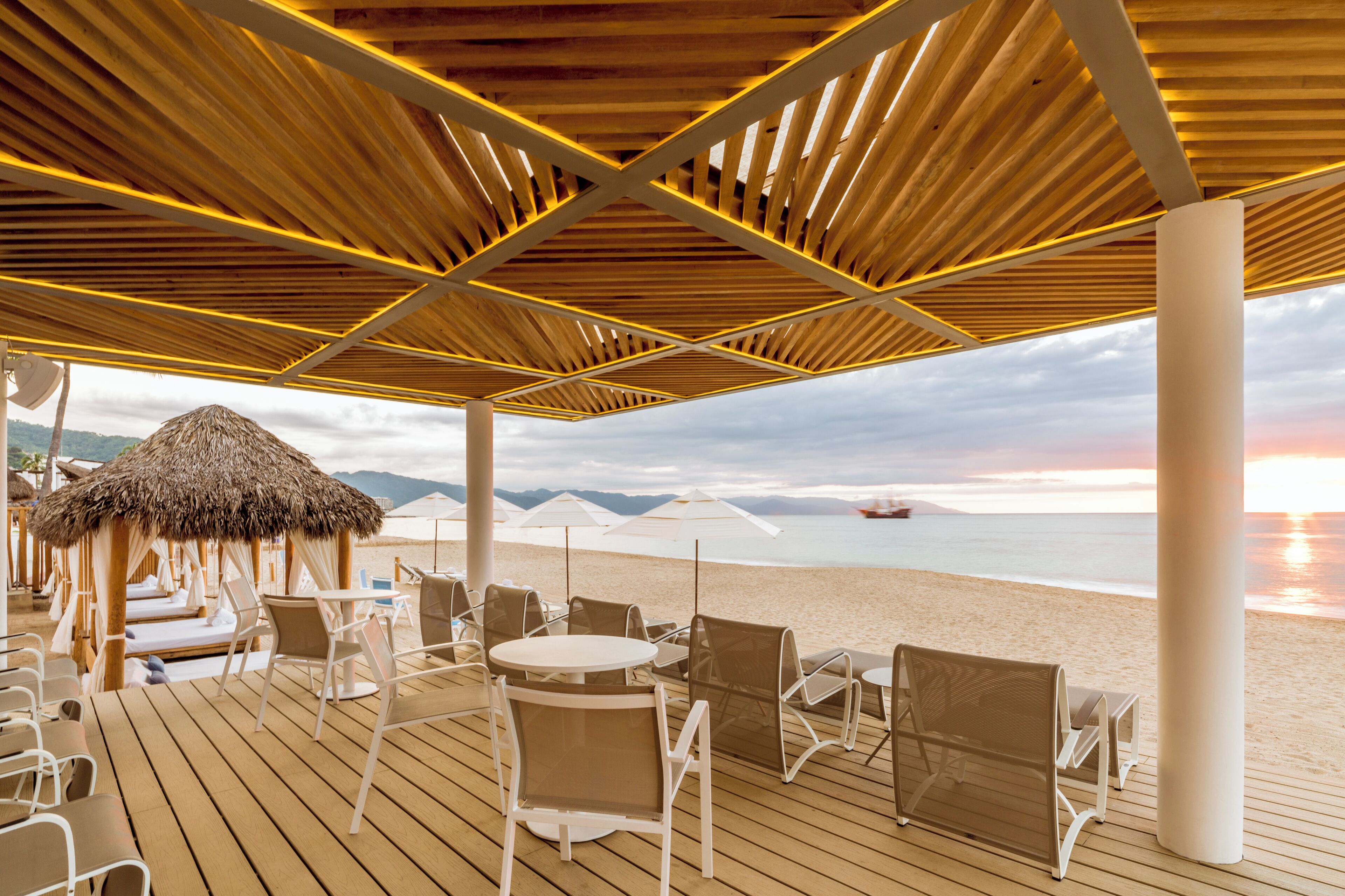 A beachside terrace with wooden flooring, white chairs, and tables, overlooking the sea and mountains.