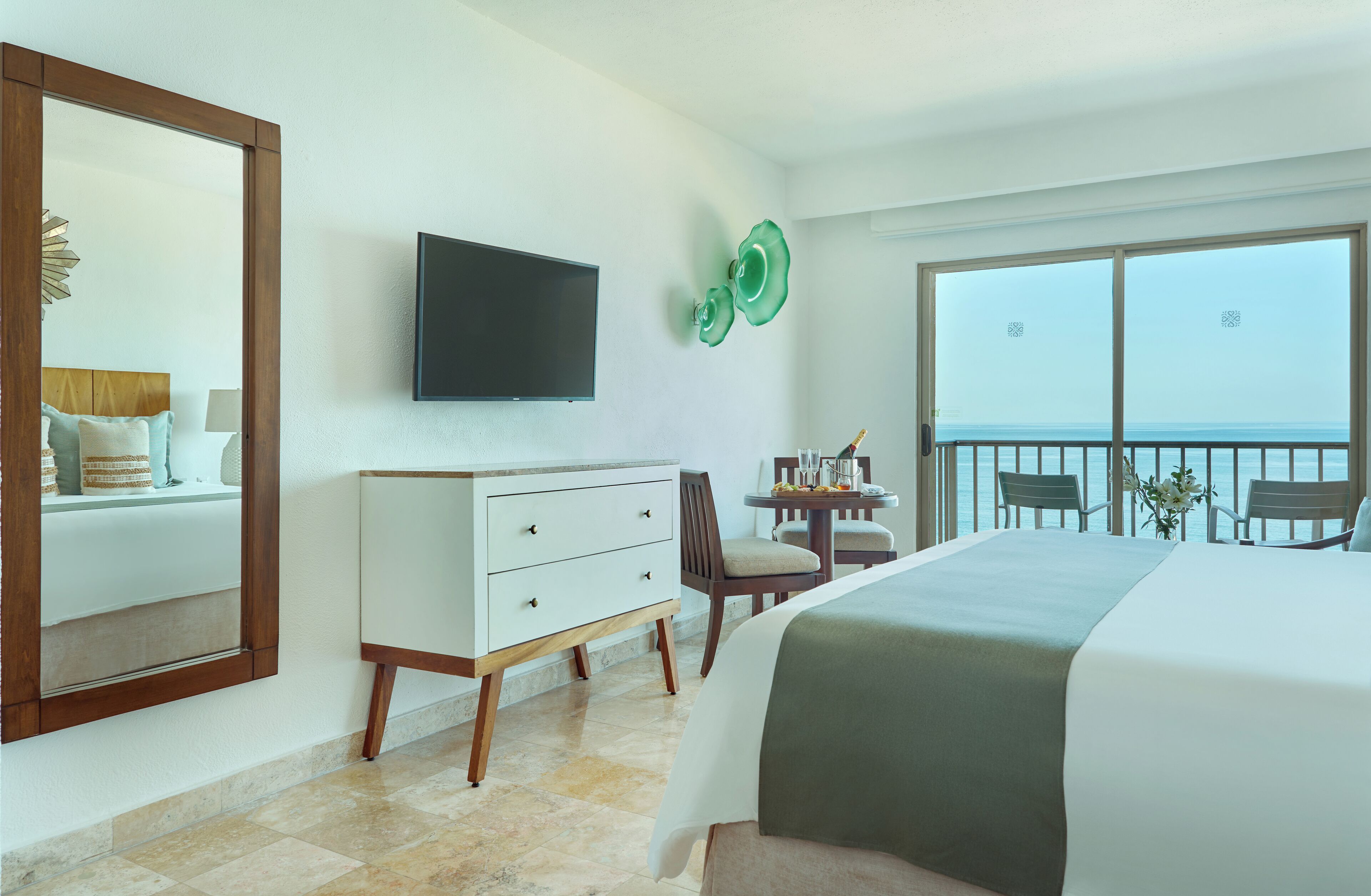 Ein Hotelzimmer mit Bett, Kommode, Fernseher, einem Balkon mit Meerblick und einem Essbereich mit Tisch und Stühlen.