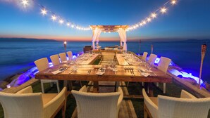 Un área de comedor al aire libre con una mesa de madera y sillas de mimbre, con vista al mar y un cielo al atardecer.