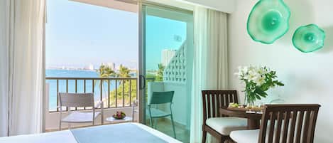 Habitación de hotel con balcón, una cama, dos sillas, una mesita con un florero y vistas a la ciudad y al mar.