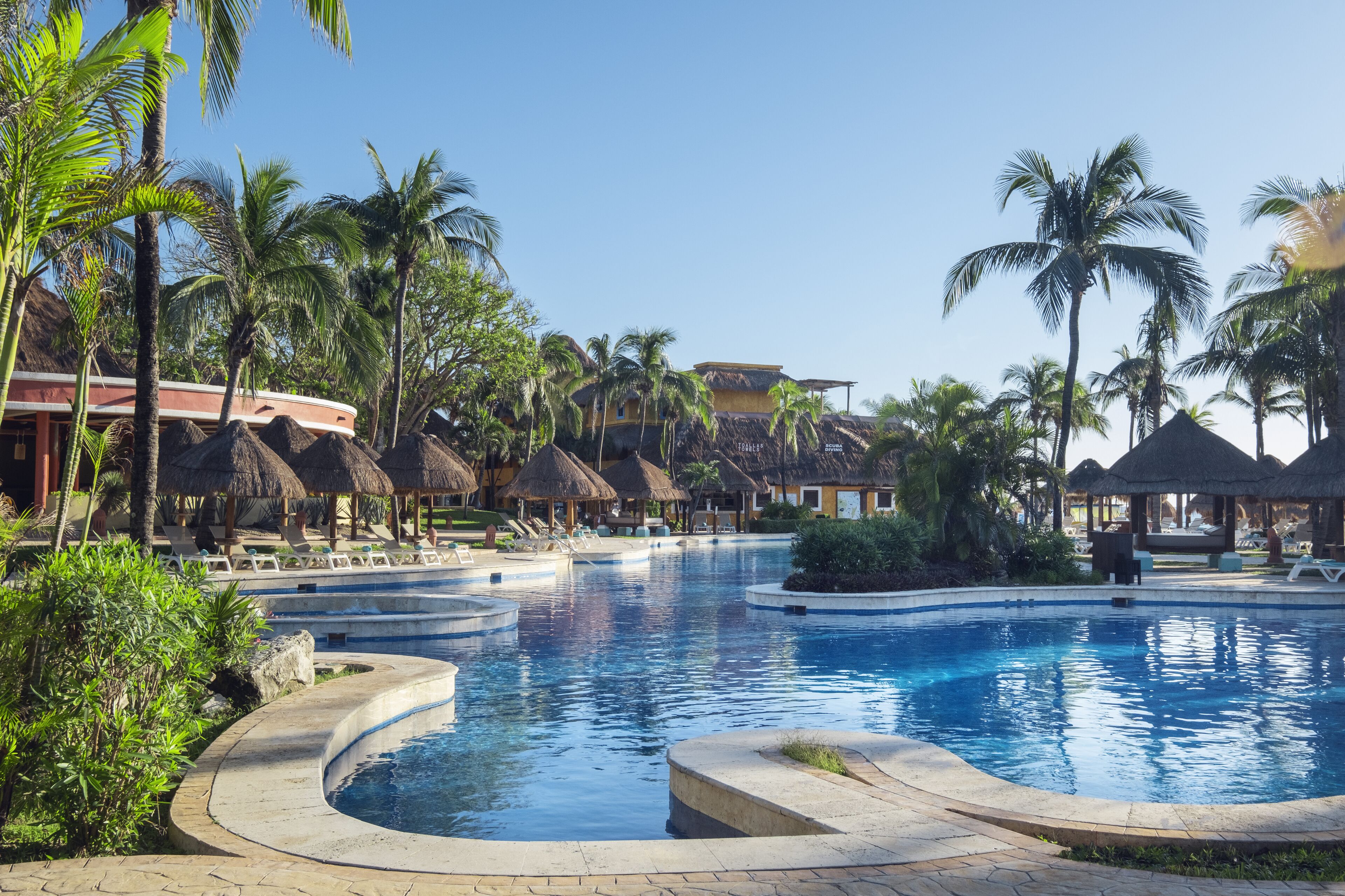 Un área de piscina en un resort con múltiples sillas de descanso y estructuras de techo de paja.