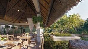 An indoor dining area with wooden tables and chairs, a thatched roof, hanging plants, and a view of a pond with rocks and foliage. - Iberostar Waves Quetzal - All Inclusive (Playa del Carmen)