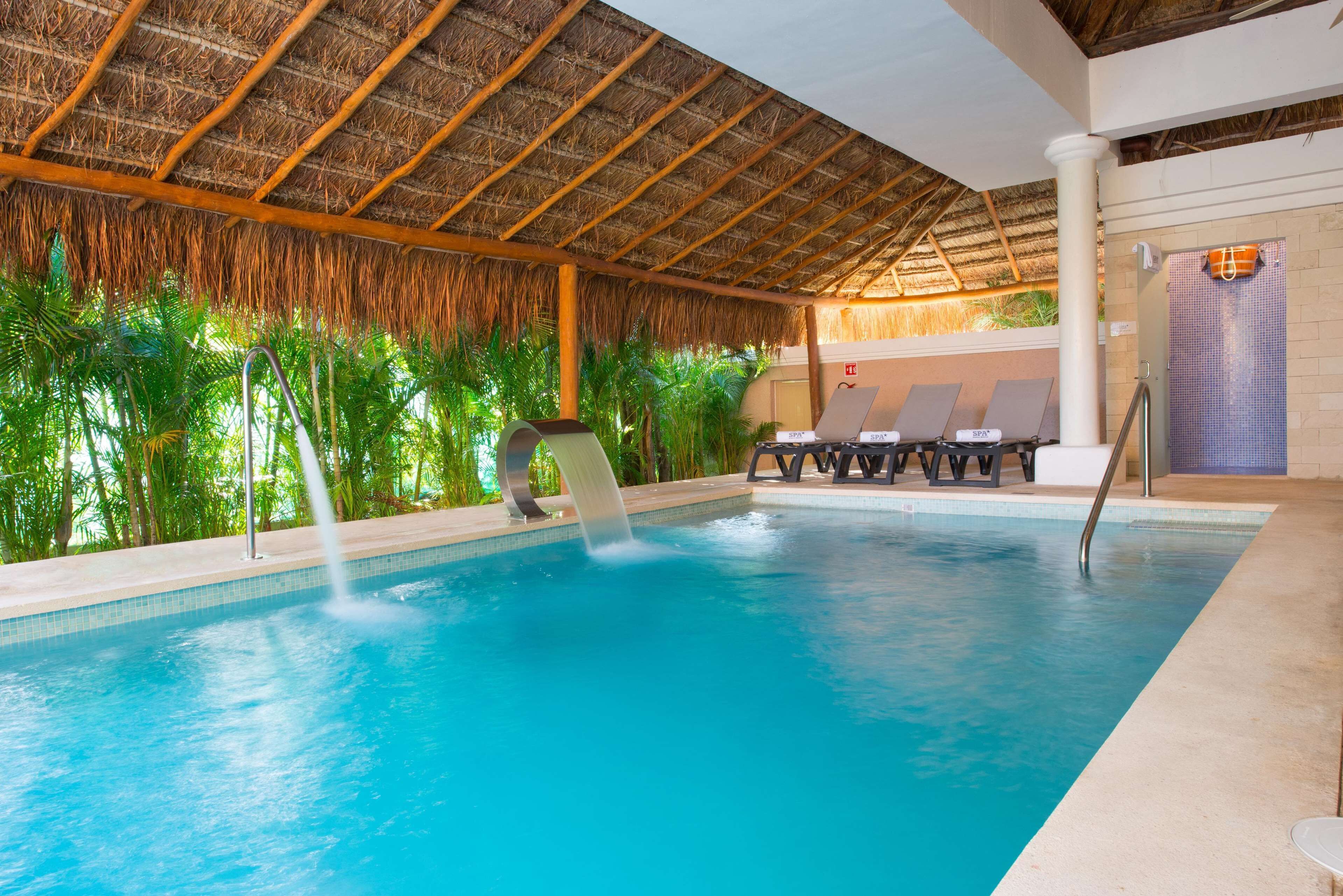 Piscina con techo de paja, sillas de descanso y vista a palmeras.