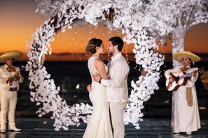 Una pareja se abraza bajo un arco floral con músicos vestidos con trajes tradicionales.
