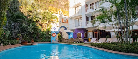 Una piscina rodeada de abundante vegetación y un edificio de varios pisos con balcones.