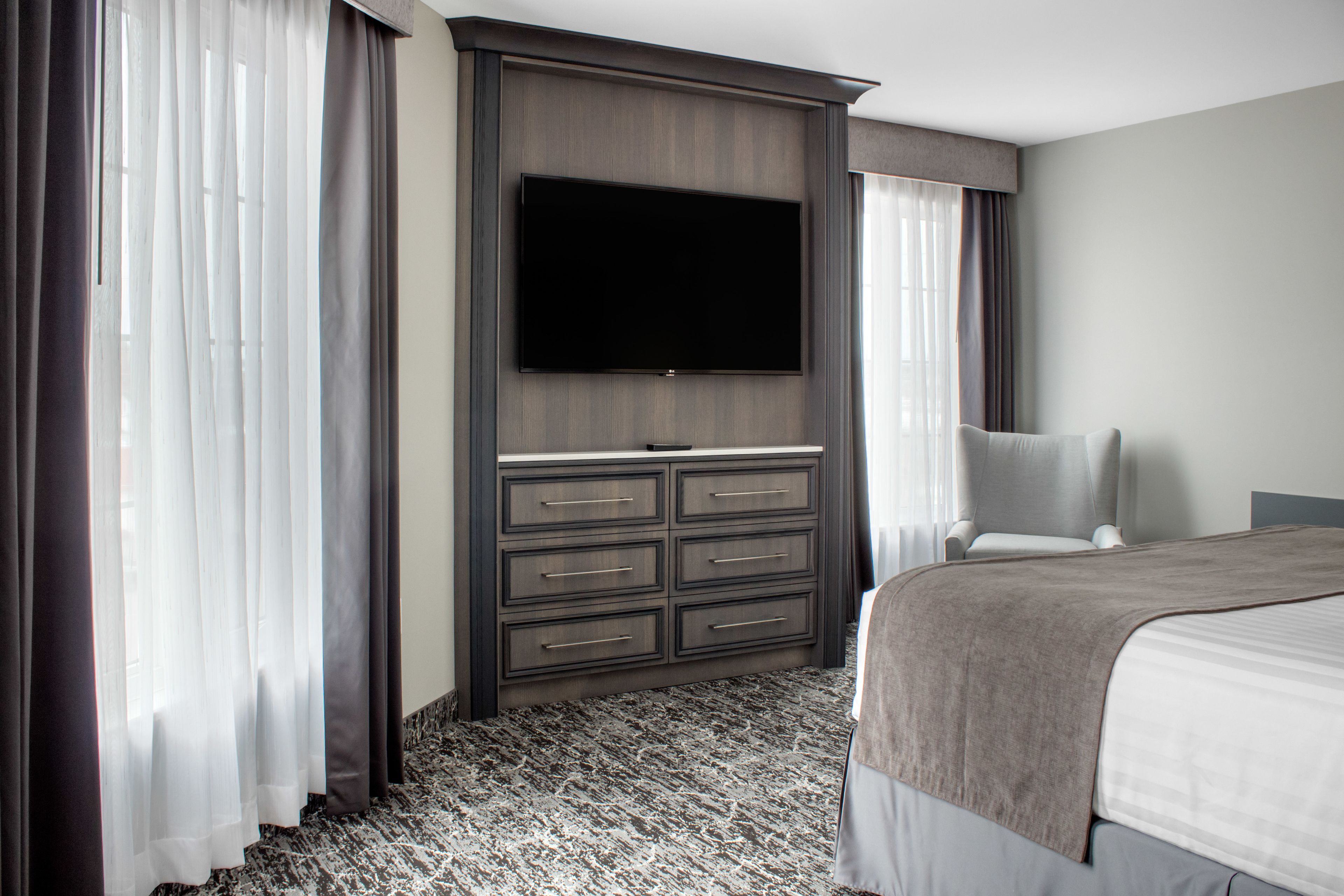 Habitación de hotel con un televisor de pantalla plana grande montado en la pared, una cama con manta gris, una silla y un escritorio con cajones.