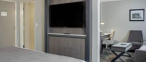 Une chambre d’hôtel avec un grand lit, un téléviseur à écran plat fixé sur un panneau en bois foncé, un bureau avec une chaise et une petite table avec une télécommande.