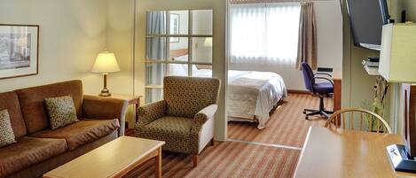 Una habitación de hotel con un sofá marrón, una mesa de madera, un sillón estampado, una cama, un televisor y una ventana con cortinas.