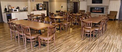 A spacious dining area with multiple wooden tables and chairs, a kitchen area with appliances, and a flat-screen TV mounted on the wall.
