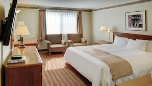 Une chambre d’hôtel avec un grand lit, deux fauteuils, un bureau avec une lampe, une télévision et un tableau encadré au mur.