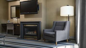 Una habitación de hotel moderna con una televisión de pantalla plana montada en la pared, chimenea, un sillón cómodo, un escritorio con lámpara y un amplio ventanal con cortinas.