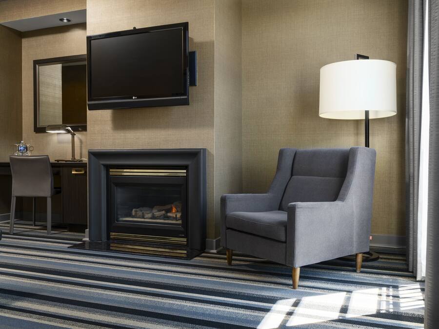 Una habitación de hotel moderna con una televisión de pantalla plana montada en la pared, chimenea, un sillón cómodo, un escritorio con lámpara y un amplio ventanal con cortinas.