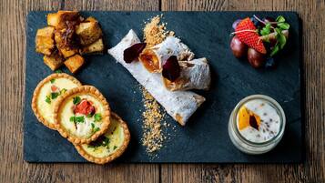 Diversos doces em uma tábua de ardósia, incluindo tartes, um bolo com açúcar em pó e um copo de sobremesa cremosa com pedaços de fruta.