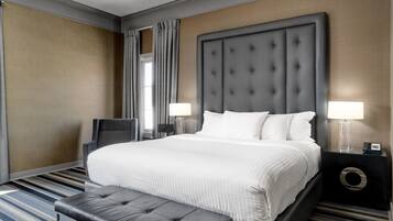 Habitación de hotel con una cama grande, un banco, dos mesitas de noche con lámparas y vistas a la ciudad desde la ventana.
