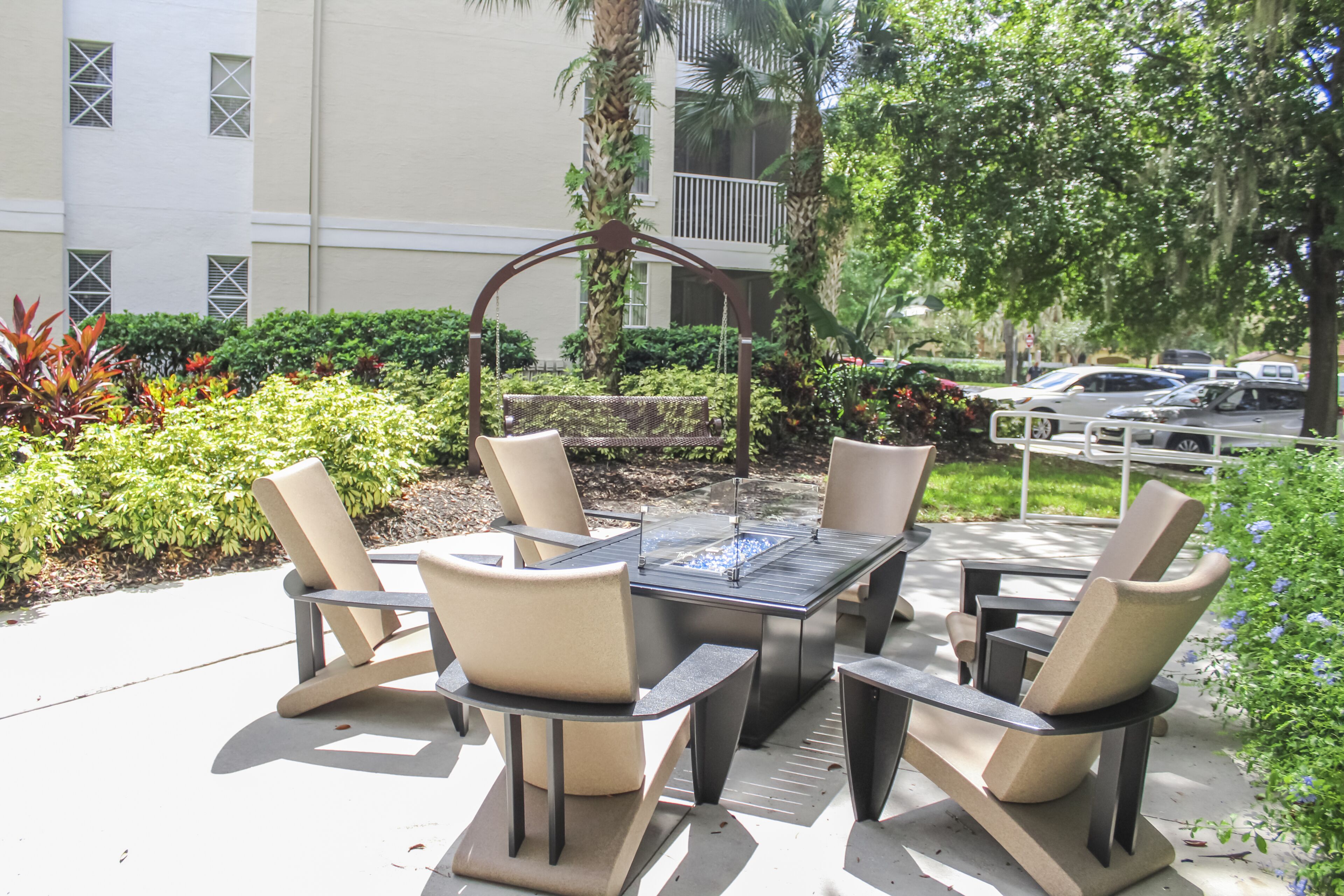 An outdoor patio with a table and chairs, surrounded by greenery and a building in the background.