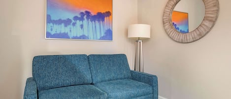 A room with a blue sofa, a painting of a sunset, a round mirror, and a floor lamp.