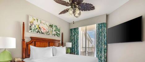 A hotel room with a large bed, a wooden headboard, a ceiling fan, a flat-screen TV, a nightstand with a lamp, and a window with curtains.
