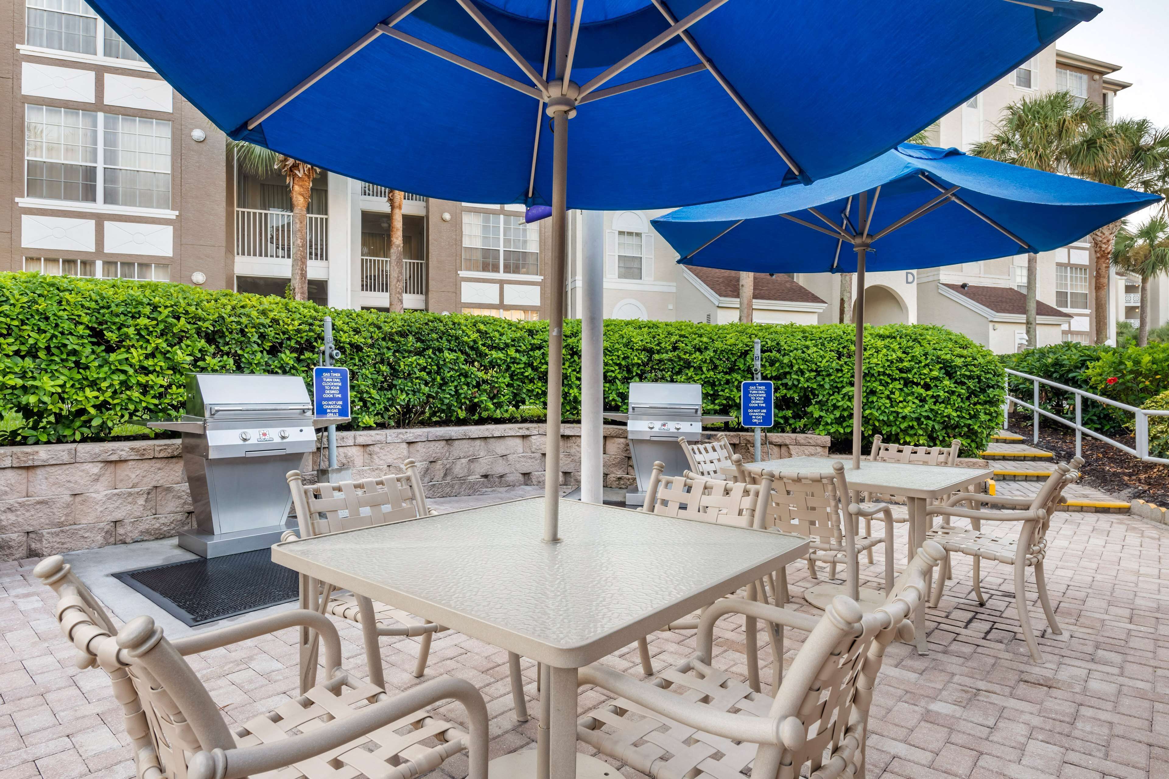 Un espace extérieur aménagé en terrasse, avec des tables et des chaises, des parasols bleus et un barbecue.
