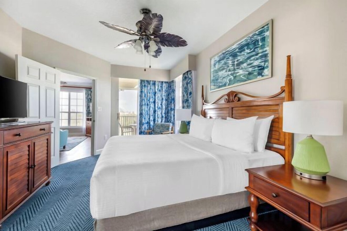 A hotel room with a bed, a wooden nightstand, a television, a ceiling fan, and a painting on the wall.