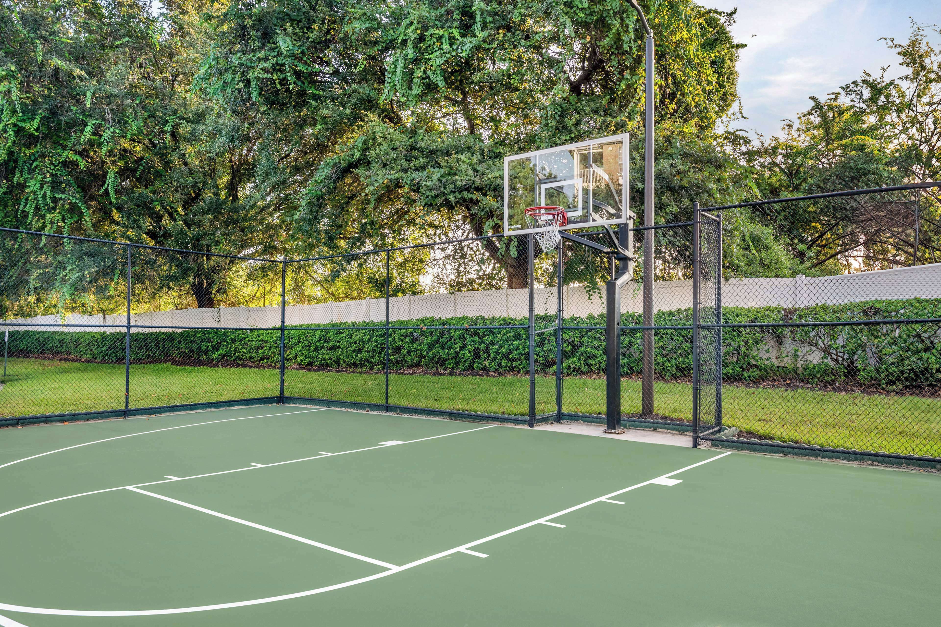 An outdoor basketball court with a chain-link fence, surrounded by trees and a hedge.