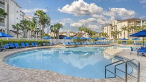 A swimming pool with blue lounge chairs and umbrellas, surrounded by palm trees and buildings.