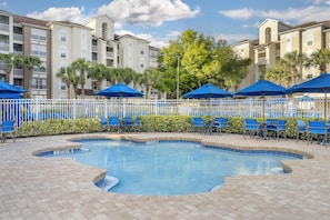 A swimming pool with blue chairs and umbrellas, surrounded by a white fence and palm trees.