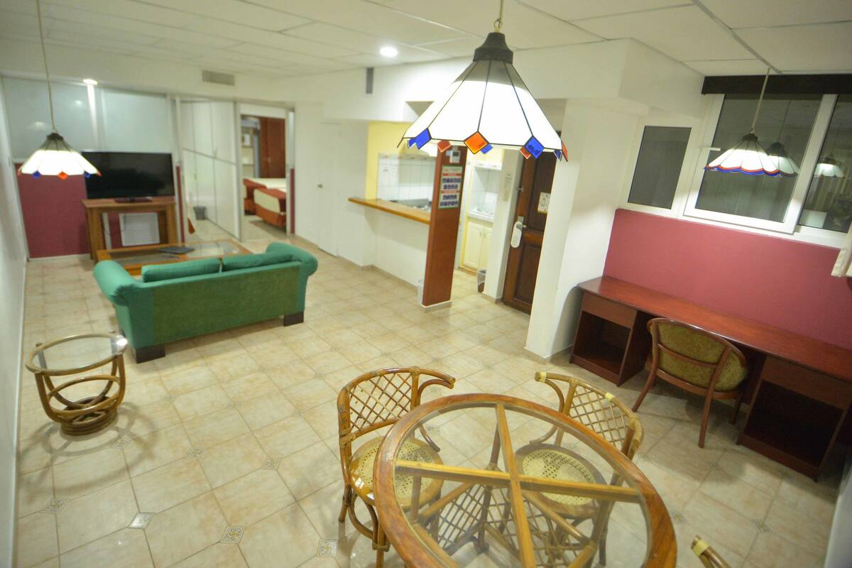 Una sala de estar con un sofá verde, televisor, mesa de comedor con sillas y una zona de cocina al fondo.