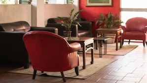 Ein Wohnzimmer mit einem roten Sessel, einem braunen Ledersofa und einem Holztisch. Auf einem Beistelltisch stehen Blumentöpfe und an der Wand hängt ein Gemälde.