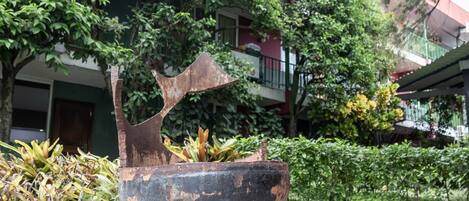 A large, rusted metal bowl in an outdoor setting with greenery and a building in the background.