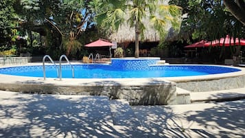 Una piscina circular con borde de piedra, rodeada de sillas de descanso y sombrillas, ubicada en un jardín tropical con árboles y una estructura de techo de paja.