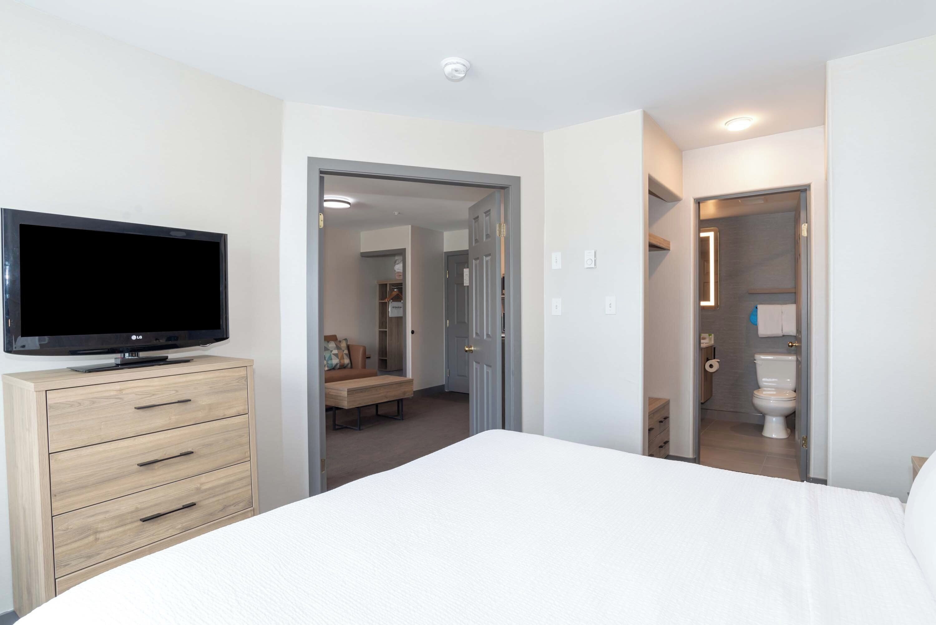 Quarto de hotel com cama, televisão sobre uma cômoda de madeira e banheiro visível através de uma porta aberta.