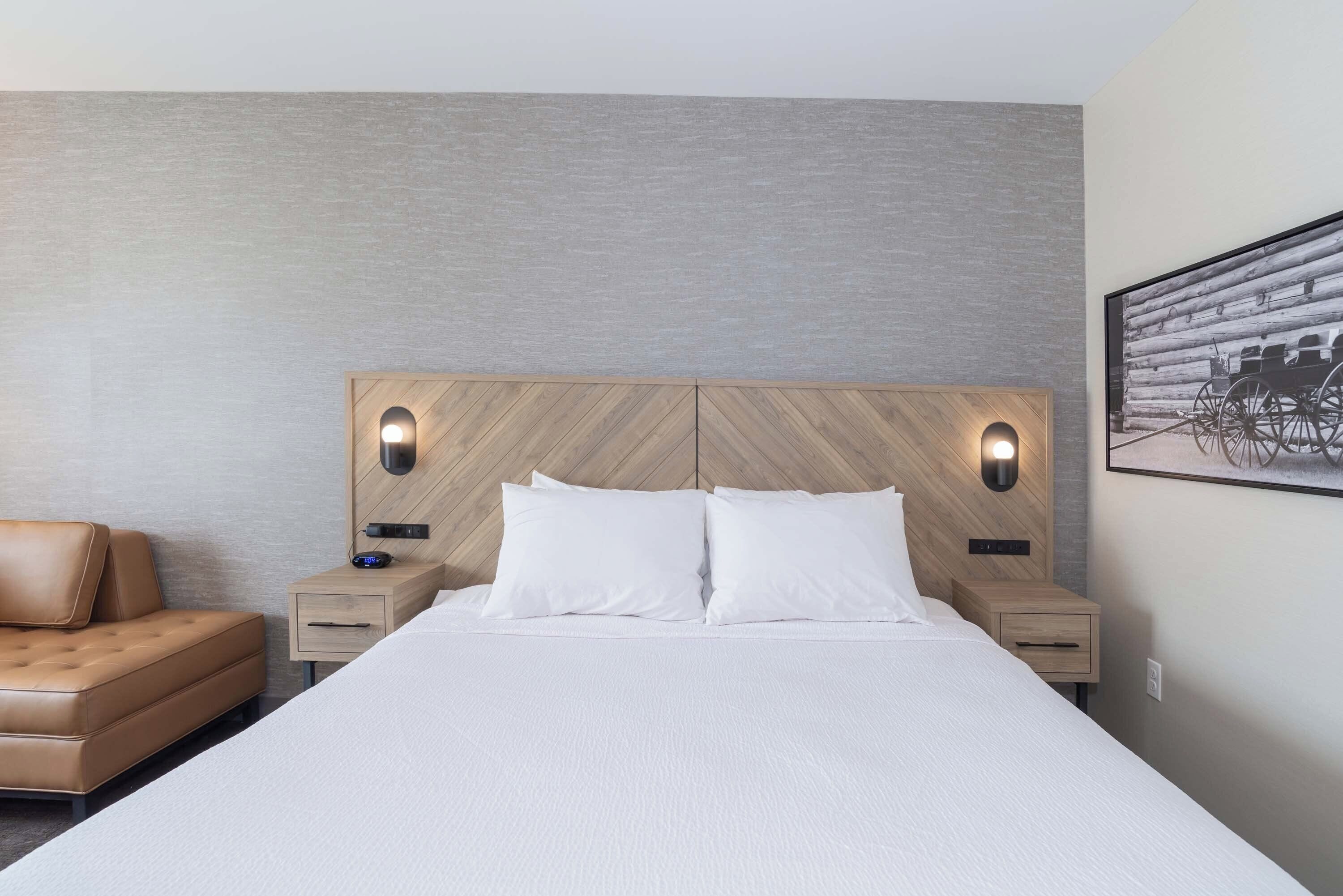 Una habitación de hotel con una cama grande, dos mesitas de noche con lámparas, un cabecero de madera, un sillón de cuero marrón y un cuadro enmarcado en la pared.