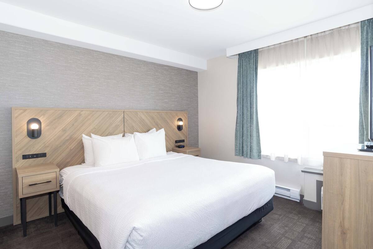 A modern hotel room with a large bed, a wooden headboard, two bedside tables, a nightstand, a desk, and a window with curtains.