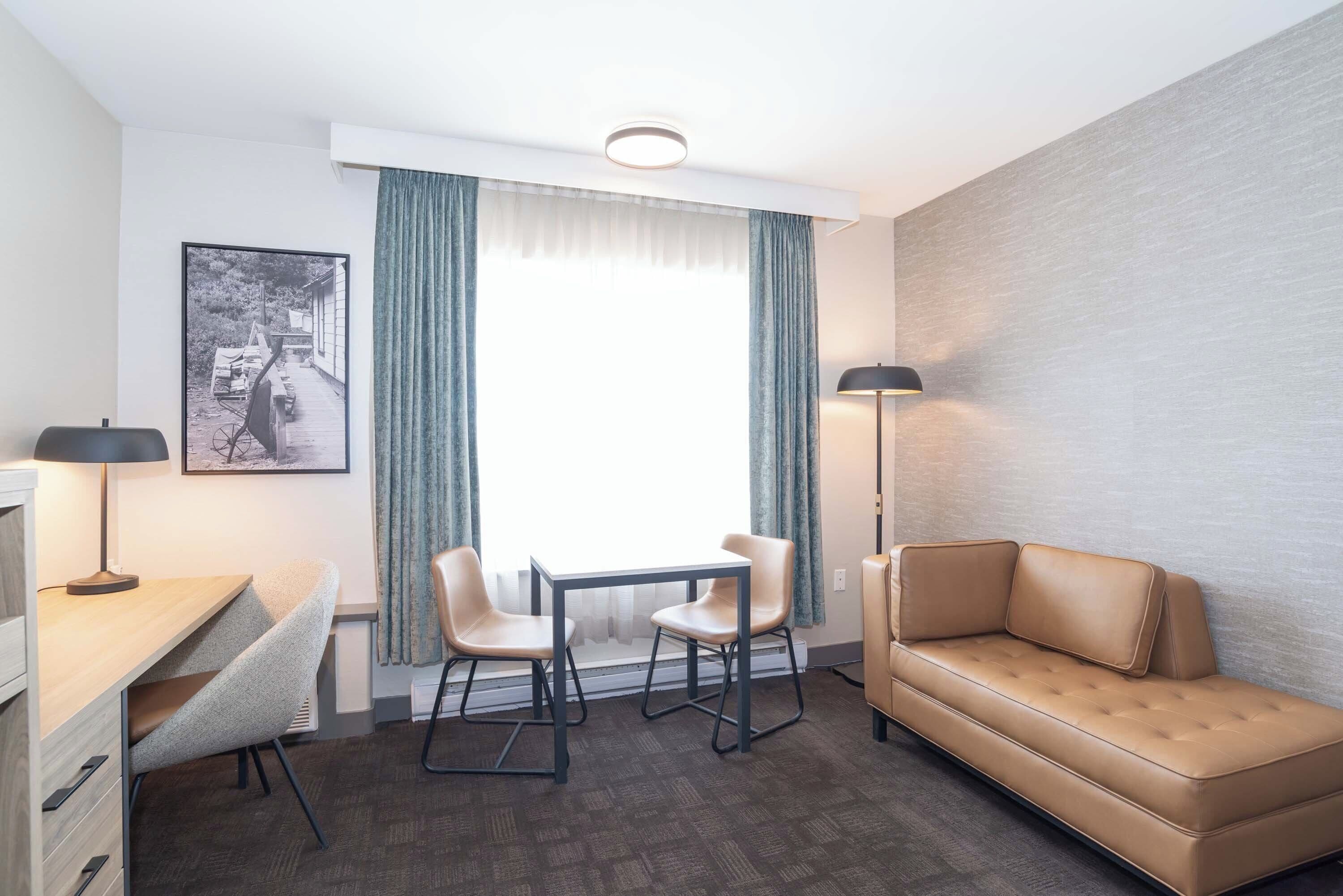 A compact hotel room with a sofa, a small table with two chairs, a desk, a lamp, and a framed picture on the wall.