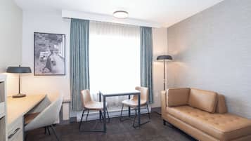A compact hotel room with a sofa, a small table with two chairs, a desk, a lamp, and a framed picture on the wall.