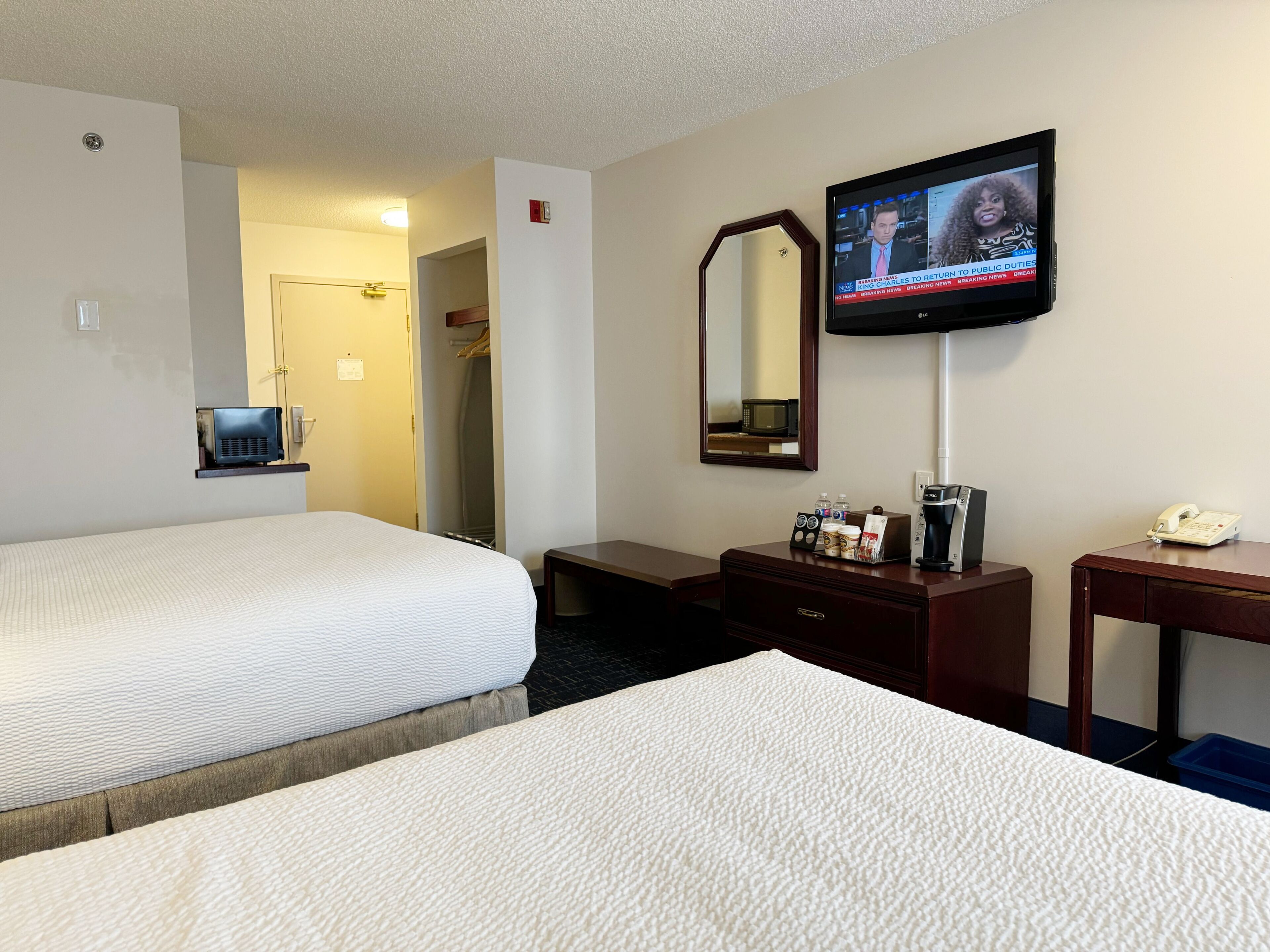 Habitación de hotel con dos camas, un televisor montado en la pared, un escritorio con teléfono y cafetera, y un espejo sobre el escritorio.