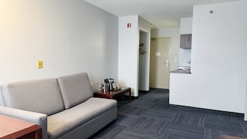 A modern hotel room with a grey sofa, a wooden side table, a kitchenette, and a hallway leading to a bathroom.