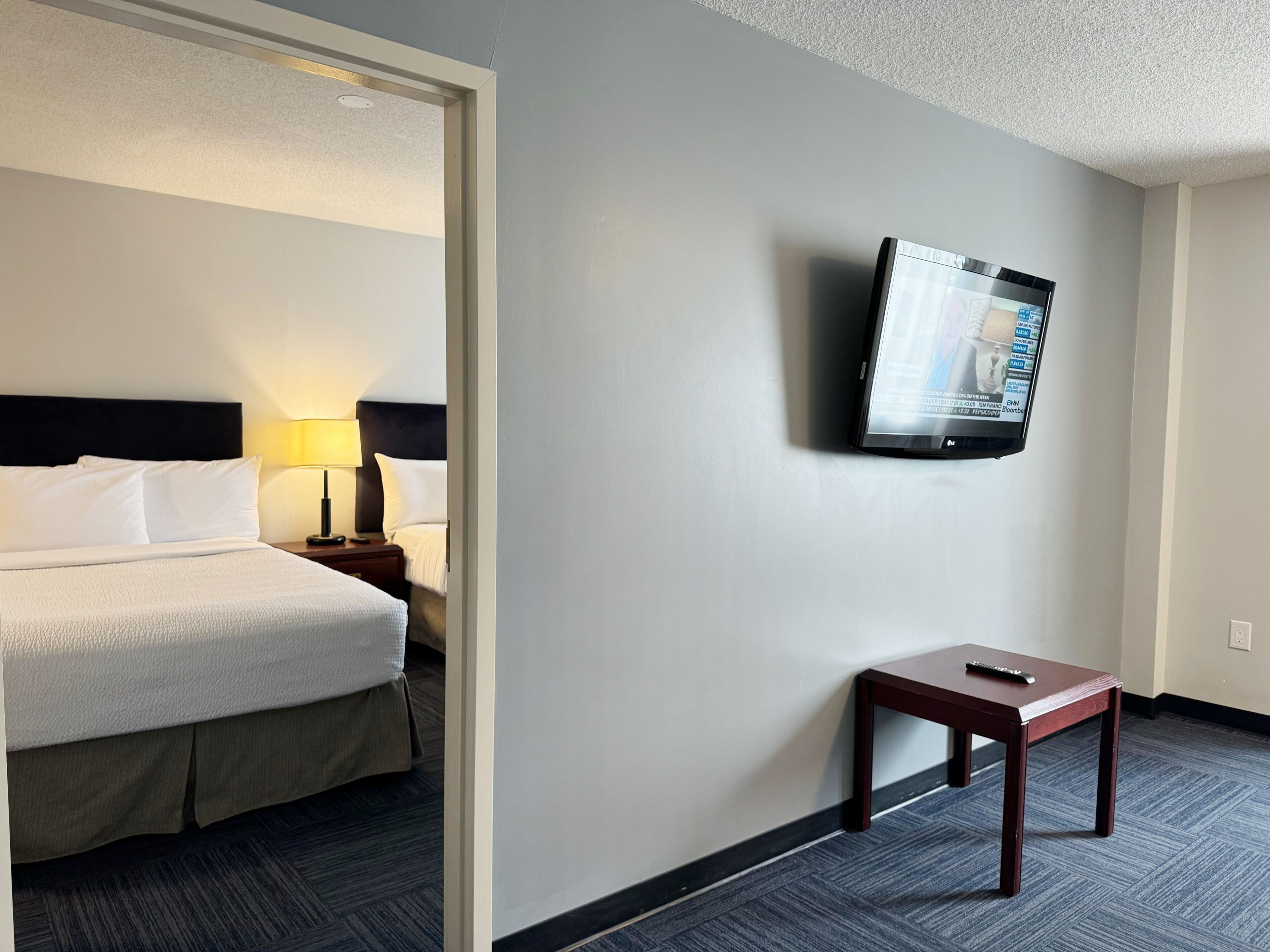 A hotel room with a bed, a nightstand with a lamp, a TV mounted on the wall, and a small table with a remote control.