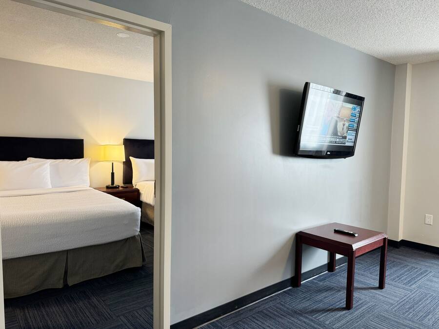 A hotel room with a bed, a nightstand with a lamp, a TV mounted on the wall, and a small table with a remote control.